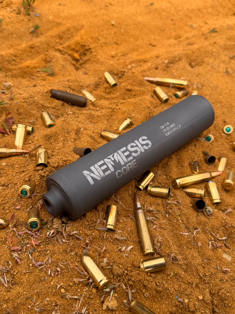 Саундмодератор NEMESIS CORE 9mm лежить на піску серед стріляних гільз - видно гравіювання NEMESIS CORE на корпусі
