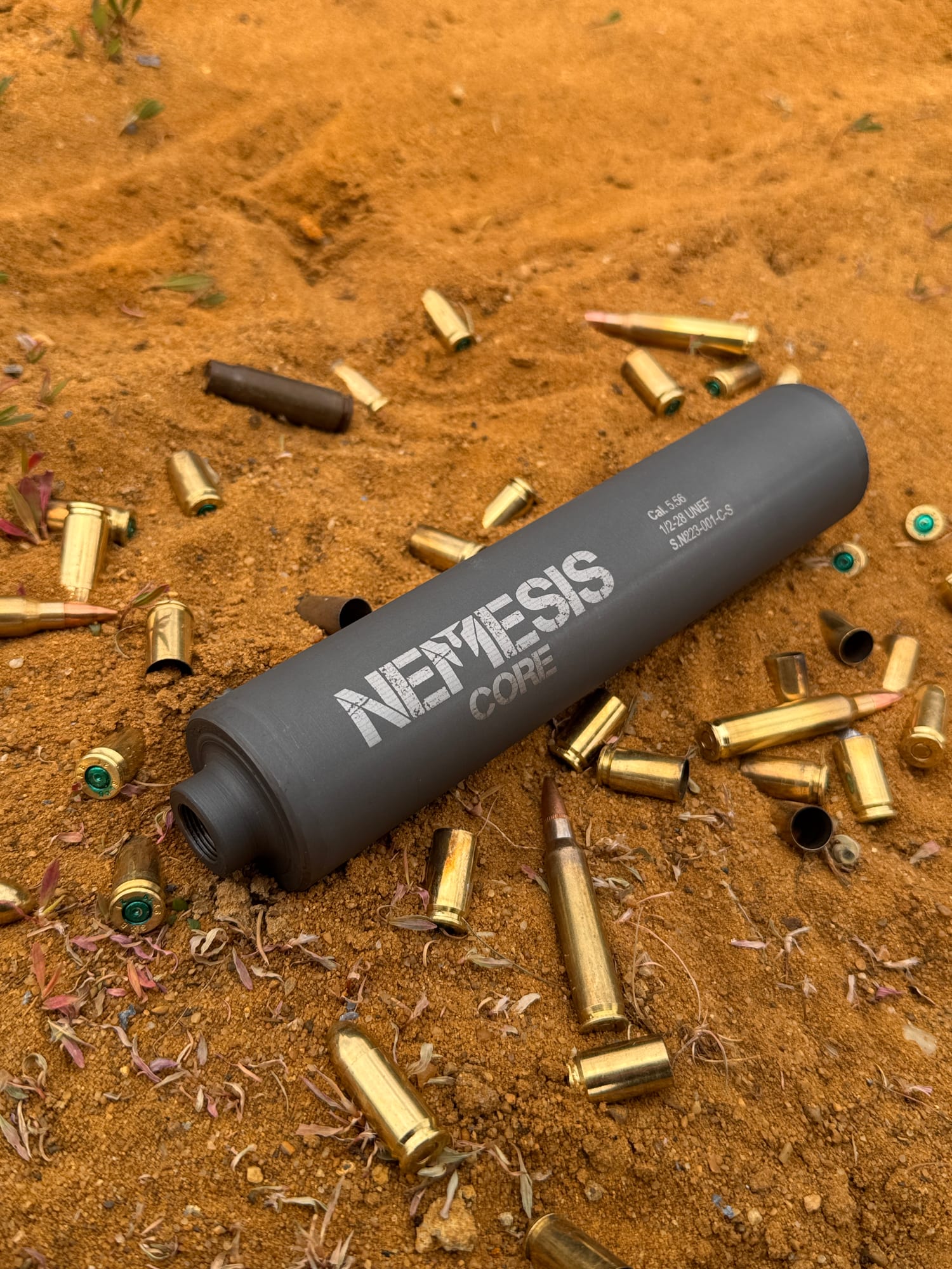Саундмодератор NEMESIS CORE 9mm лежить на піску серед стріляних гільз - видно гравіювання NEMESIS CORE на корпусі