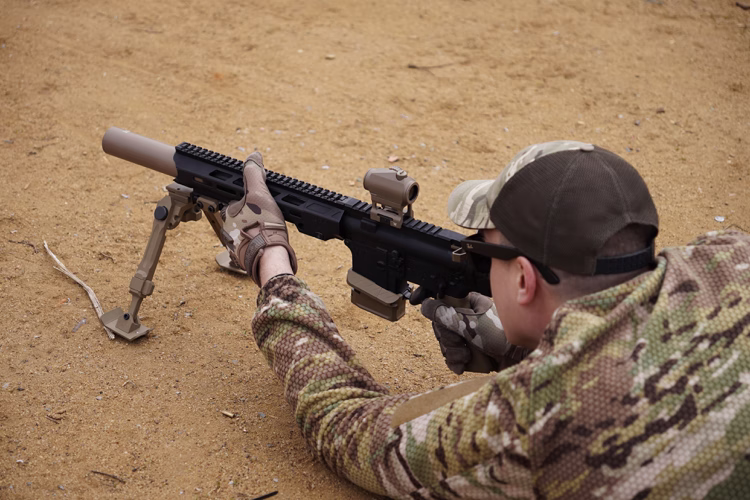 Стрілець у мультикамі цілиться з AR-15 5.56 з глушником NEMESIS CORE на сошках, вид збоку крупним планом