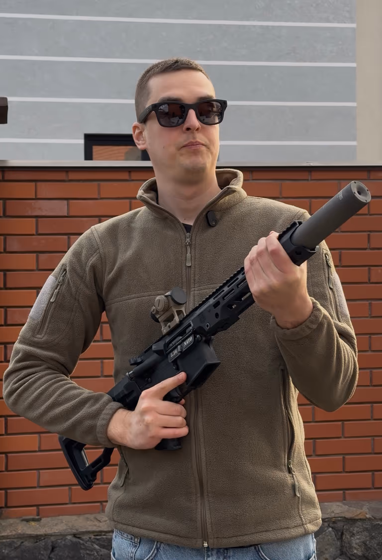 Стрілець тримає PCC із глушником NEMESIS CORE 9mm чорним, вид у повний ріст на тлі цегляної стіни