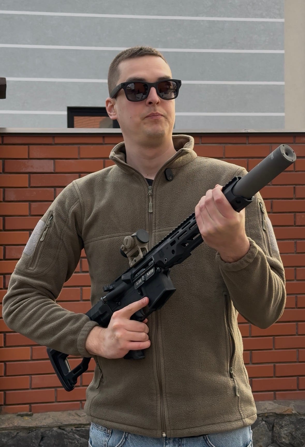 Стрілець тримає PCC із глушником NEMESIS CORE 9mm чорним, вид у повний ріст на тлі цегляної стіни